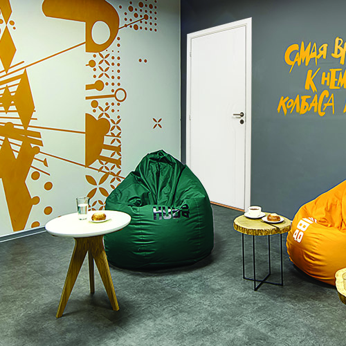 Canteen in a coworking. Left wall is white with orange images. Right wall is gray with orange inscription and the white door on the left. There are three small round tables with tea, water and pies. There are also two green and one orange poufs for sitting. On the floor there are gray tiles. At the top there are communications and lamps.